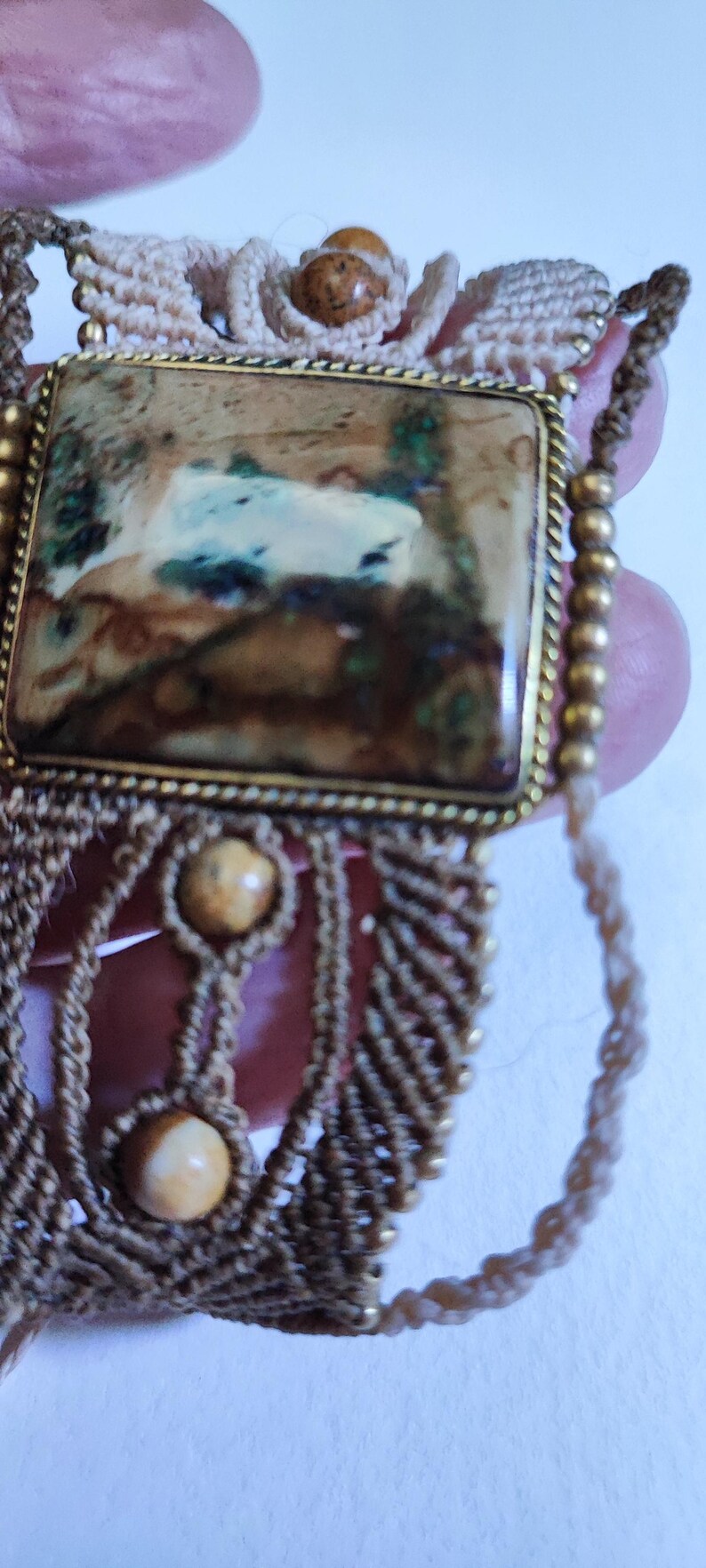 May include: Close-up of a handcrafted macrame bracelet featuring a rectangular stone with brown, green, and white hues set in a gold-colored frame. The bracelet has a beige macrame design with two beige beads.