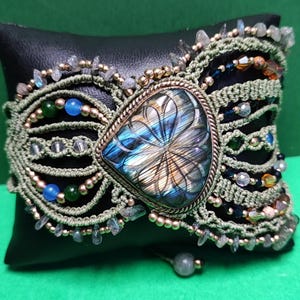 Puede incluir: Una pulsera de macramé con una gran piedra de labradorita tallada en forma triangular en el centro. La banda está hecha de cordón verde oliva, adornada con pequeñas cuentas metálicas y varias cuentas de vidrio de colores azul, verde y ámbar.