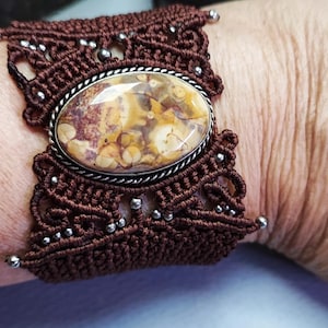 Può includere: Bracciale in macramè marrone con un elemento centrale in pietra ovale e screziata. La pietra è incastonata in una lunetta color argento e circondata da intricati nodi macramè. Piccole perline argentate accentuano il design macramè.