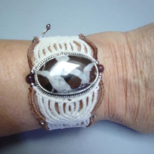 Può includere: Un braccialetto in macramé bianco e marrone con un ciondolo ovale argentato con una pietra marrone e bianca.