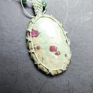 Op de afbeelding: Ovale hangerketting met een lichtgroene steen met rode insluitsels, gevat in een macrame-net. De ketting heeft een wit koord met groene en bruine kralen.