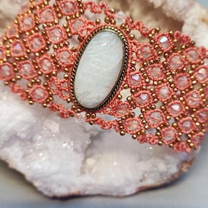 May include: A handmade bracelet featuring a central oval-shaped white stone set in a bronze-colored metal frame. The bracelet has a coral-colored, open-weave design with small gold-colored beads and clear crystals. The bracelet is displayed on a white, crystalline surface.