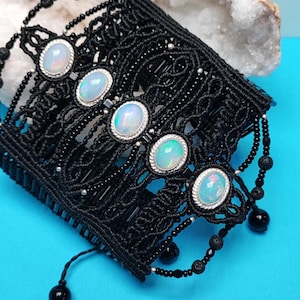 May include: A black macrame cuff bracelet featuring five oval-shaped iridescent opals set in silver-toned bezels. The intricate black cord weave is accented with small black beads and dangling black bead strands, creating a detailed texture.