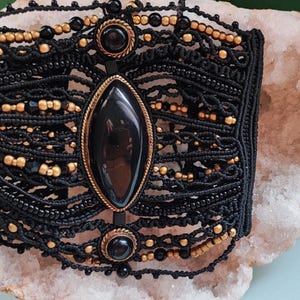 May include: Black macramé bracelet with gold-coloured beads and accents. The bracelet features a large, oval, black stone in the centre, framed by a gold-coloured setting. Additional black stones and gold beads are incorporated.