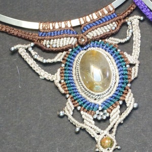 May include: A beige macrame necklace with a silver metal base. The necklace features a large, oval-shaped, clear quartz stone surrounded by blue, green, and brown macrame. The necklace also has smaller, round, silver beads and a smaller, green stone at the bottom.