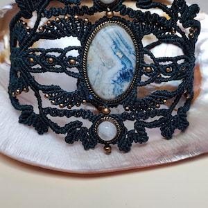 Peut inclure: Un bracelet en macramé bleu foncé avec une pièce maîtresse ovale en pierre marbrée bleue et blanche. Le bracelet comporte de petites perles dorées et deux pierres blanches rondes. Le design est complexe et détaillé.