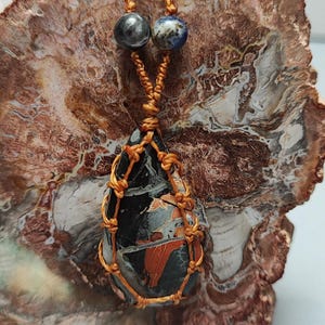 May include: A teardrop-shaped pendant necklace featuring a black, red, and gray stone encased in a woven orange cord. The necklace has two small, round beads at the top. The pendant is displayed on a brown and white patterned surface.