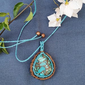 May include: A handcrafted pendant necklace featuring a teardrop-shaped turquoise stone with brown accents, encased in a brown and turquoise macrame setting. The necklace has a turquoise cord and copper beads. The background is a blue fabric.