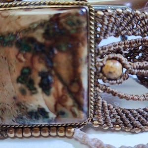 May include: A handcrafted macrame bracelet featuring a large rectangular stone with brown, green, and beige patterns, set in a gold-toned frame. The bracelet incorporates woven macrame detailing and small, round beads, creating a bohemian aesthetic.