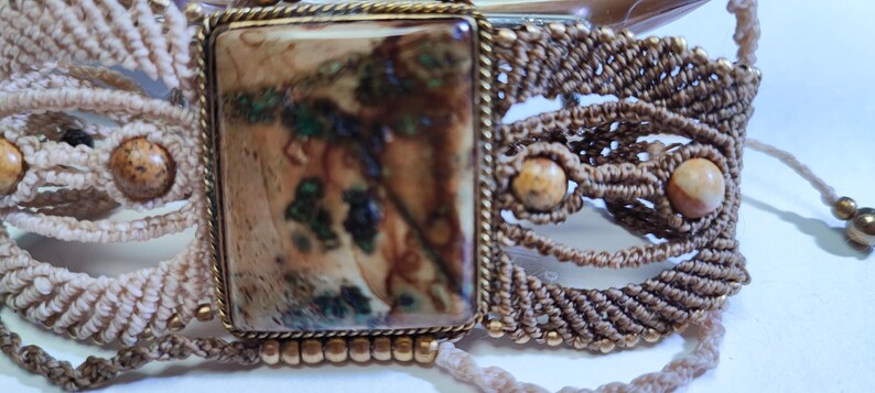 May include: A handcrafted macrame bracelet featuring a large rectangular stone with brown, green, and beige patterns, set in a gold-toned frame. The bracelet incorporates woven macrame detailing and small, round beads, creating a bohemian aesthetic.