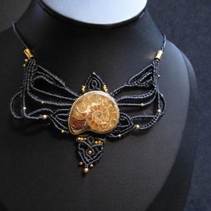 May include: A black macrame necklace with a gold-toned ammonite fossil pendant. The necklace is adorned with small gold beads.