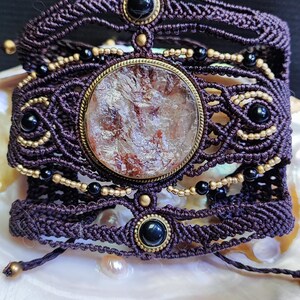 Puede incluir: Pulsera de macramé artesanal en morado oscuro, con una gran pieza central de piedra redonda y multicolor. Acentuada con cuentas negras y doradas y cabujones negros. La pulsera se exhibe sobre una superficie blanca.