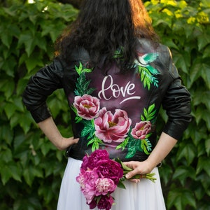 Puede incluir: Una chaqueta de cuero negra con un dise&ntilde;o floral y un p&aacute;jaro pintado en la espalda. La chaqueta tiene la palabra "love" escrita en pintura blanca. La persona sostiene un ramo de peon&iacute;as rosas.