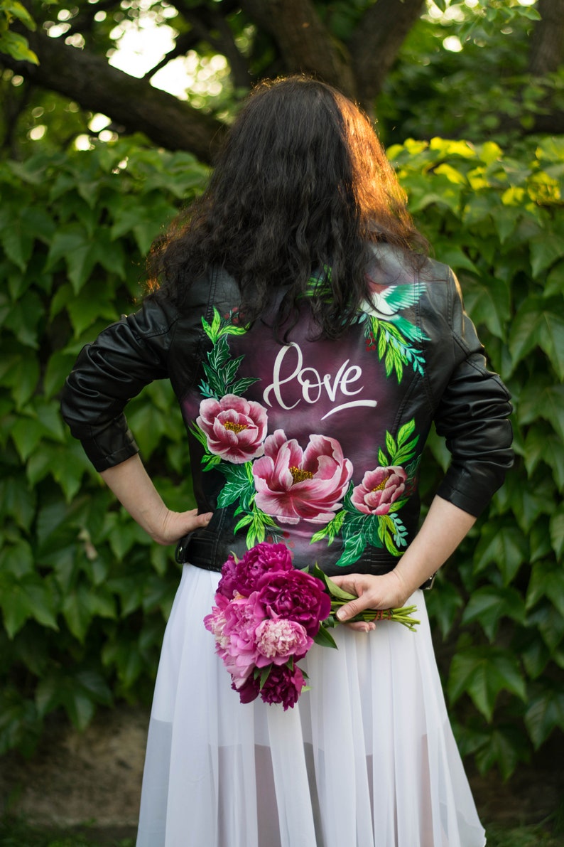 Puede incluir: Una chaqueta de cuero negra con un dise&ntilde;o floral y un p&aacute;jaro pintado en la espalda. La chaqueta tiene la palabra "love" escrita en pintura blanca. La persona sostiene un ramo de peon&iacute;as rosas.