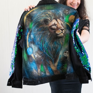 Könnte beinhalten: Eine schwarze Jeansjacke mit einem handgemalten Löwenmotiv auf der Rückseite. Der Löwe ist von einem galaktischen Hintergrund mit Sternen und Planeten umgeben. Die Jacke hat grüne und blaue Pailletten an den Ärmeln.
