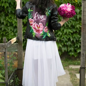 Puede incluir: Una chaqueta de cuero negra con un dise&ntilde;o floral y la palabra "love" pintada en la espalda. La chaqueta se lleva sobre una falda blanca.
