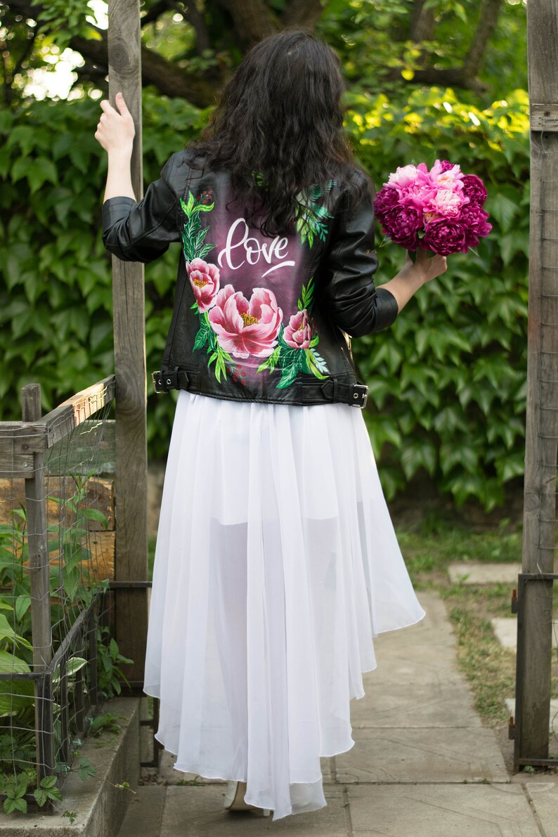 Puede incluir: Una chaqueta de cuero negra con un dise&ntilde;o floral y la palabra "love" pintada en la espalda. La chaqueta se lleva sobre una falda blanca.