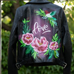 Puede incluir: Chaqueta de cuero negra con un dise&ntilde;o floral pintado en la espalda. El dise&ntilde;o incluye flores rosas, hojas verdes y la palabra "love" en letras blancas.