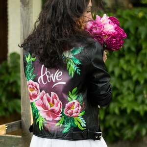 Puede incluir: Una chaqueta de cuero negra con un dise&ntilde;o floral pintado en la espalda. El dise&ntilde;o incluye flores rosas, hojas verdes y la palabra "love" en letras blancas.