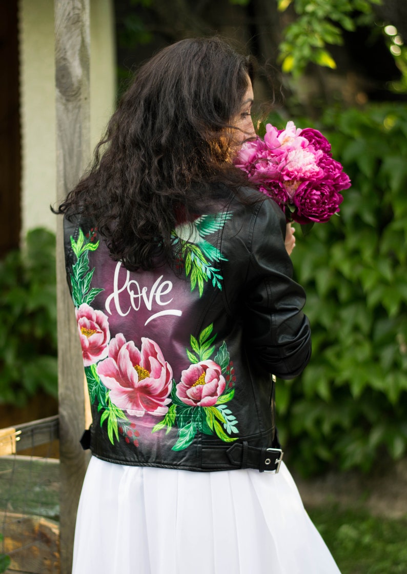 Puede incluir: Una chaqueta de cuero negra con un dise&ntilde;o floral pintado en la espalda. El dise&ntilde;o incluye flores rosas, hojas verdes y la palabra "love" en letras blancas.