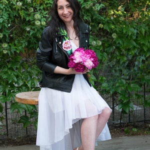 Puede incluir: Una mujer con un vestido blanco y una chaqueta de cuero negra con bordados florales sostiene un ramo de peon&iacute;as rosas. Est&aacute; sentada en un taburete de madera frente a un fondo verde frondoso.