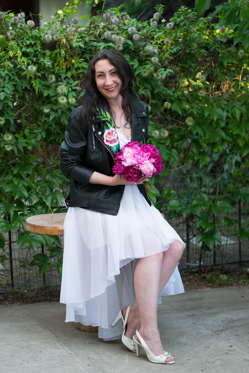 Puede incluir: Una mujer con un vestido blanco y una chaqueta de cuero negra con bordados florales sostiene un ramo de peon&iacute;as rosas. Est&aacute; sentada en un taburete de madera frente a un fondo verde frondoso.