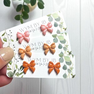 May include: Three pairs of bow-shaped stud earrings displayed on white cards with green leaf illustrations and the text WYM BY JB. The top pair is shimmering light pink, the middle pair is matte peach, and the bottom pair is glossy orange.