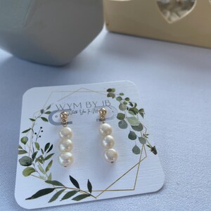 Puede incluir: Un par de pendientes de oro con tres perlas blancas colgando de cada pendiente. Los pendientes se muestran en una tarjeta blanca con follaje verde.