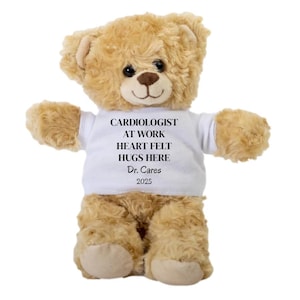 May include: A plush teddy bear with light brown fur. The bear wears a white t-shirt with the text "CARDIOLOGIST AT WORK HEART FELT HUGS HERE Dr. Cares 2025". The bear has a friendly expression and outstretched arms.