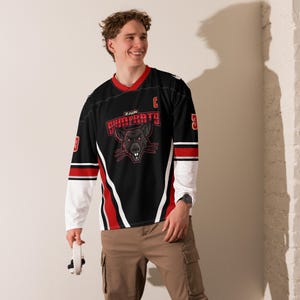 Op de afbeelding: Een zwart en rood hockeytrui met een wit en rood streepontwerp. De trui heeft een zwart rattenmasker met rode ogen en de tekst "ZAIN SHIMPRATS" op de voorkant. De trui heeft een "C" op de rechter schouder en het nummer "5" op de rechter mouw.
