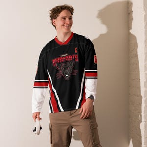 Op de afbeelding: Zwart en rood hockey-shirt met de tekst "ZAUN DUMPRATS" en een rattenafbeelding. Het shirt heeft witte mouwen en rode accenten. Het nummer "6" staat op de rechter mouw.