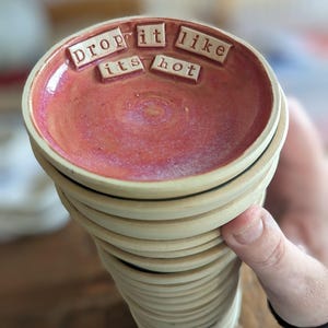 May include: A stack of round ceramic dishes with a glossy pink interior and a matte beige exterior. The top dish features the text "drop it like its hot" in raised, individual square letter tiles arranged in two rows across the centre.