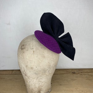 Op de afbeelding: Een paarse en zwarte wollen hoed met een grote strik. De hoed zit op een wit mannequin hoofd.