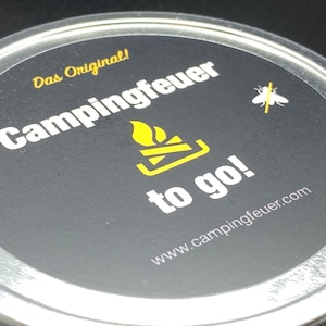 Może przedstawiać: Okrągły srebrny pojemnik z czarną etykietą, na której widnieje napis "Das Original! Campingfeuer to go! www.campingfeuer.com". Etykieta zawiera również żółty grafik płomienia i małą białą muchę z żółtą linią przekreślającą ją.