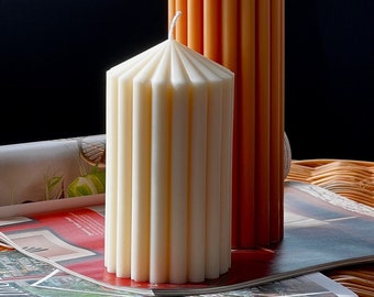 May include: Two pillar candles, one white and one orange, with a fluted design. The candles are sitting on a magazine on a wicker surface.
