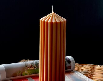 May include: A tall, orange pillar candle with a fluted design. The candle is sitting on top of a stack of magazines on a wicker surface.