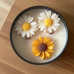 May include: A black candle jar with three white and yellow wax flowers on top. The flowers are a daisy and a sunflower.