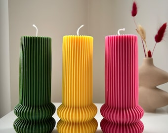 May include: Three tall, cylindrical candles with a ribbed design. The candles are green, yellow, and pink. The candles are on a white surface.
