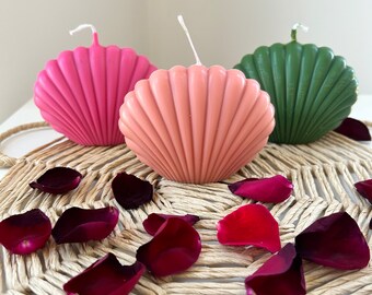 Puede incluir: Tres velas decorativas con forma de concha marina, una rosa, una melocotón y una verde, están dispuestas sobre una estera de paja tejida con pétalos de rosa roja esparcidos alrededor.