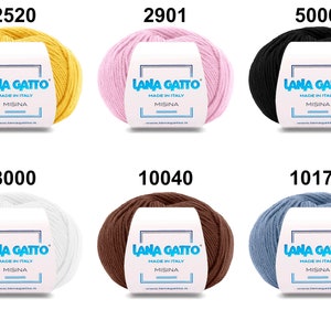 Op de afbeelding: Zes bollen garen in verschillende kleuren, waaronder geel, roze, zwart, wit, bruin en blauw. Elke bol heeft een label met de tekst "Lana Gatto" en "Made in Italy".