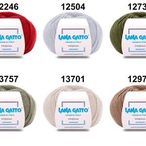 Op de afbeelding: Zes bollen garen in verschillende kleuren, waaronder rood, wit, groen, grijs, beige en bruin. Elke bol heeft een label met de tekst "Lana Gatto" en "Made in Italy".