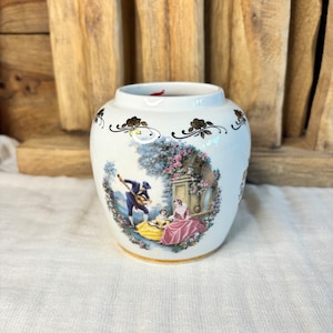 May include: White ceramic vase with a gold trim and a painted scene of a couple in a garden. The scene includes a man playing a guitar and a woman in a pink dress.