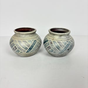 May include: Two small, round ceramic vases with a light beige base color. Each vase features a geometric pattern in shades of blue, gray, and purple. The interior of each vase is a dark brown color.