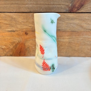 May include: A white ceramic pitcher with a spout, featuring hand-painted red fern and green insect designs. The pitcher has a unique, textured shape and is adorned with green and yellow abstract lines.