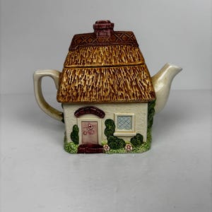 May include: A cream-coloured ceramic teapot in the shape of a cottage. It features a thatched roof, brick chimney, door, window, and decorative greenery. The teapot has brown and green accents, and is designed to resemble a quaint home.