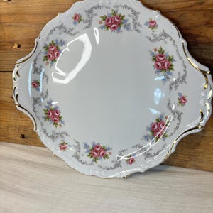 Puede incluir: Un plato de servir de porcelana blanca con borde festoneado y ribete dorado. El plato está decorado con un patrón floral de rosas rosas y azules y hojas grises.