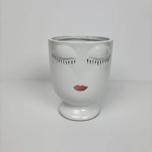 May include: White ceramic planter with a face design. The planter features closed eyes with black eyelashes and red lips. The planter is a decorative item for indoor plants, adding a touch of artistic flair to any space.