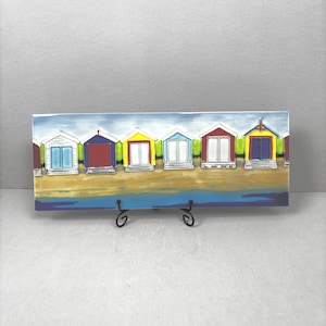 May include: A rectangular painting showing six colourful beach huts on a sandy shore. The huts have white, red, yellow, and blue doors and roofs. The artwork is displayed on a black metal stand.