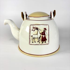 May include: Cream ceramic teapot with a Christmas design. The teapot has a square image of Santa Claus and a reindeer. The lid and handles are light brown. The teapot has a spout and a round base. The teapot is a festive item.