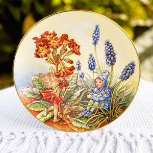 May include: A decorative ceramic plate featuring a whimsical illustration of a fairy with wings and a blue-clad gnome sitting amongst a cluster of red and pink primrose flowers and blue grape hyacinth flowers.